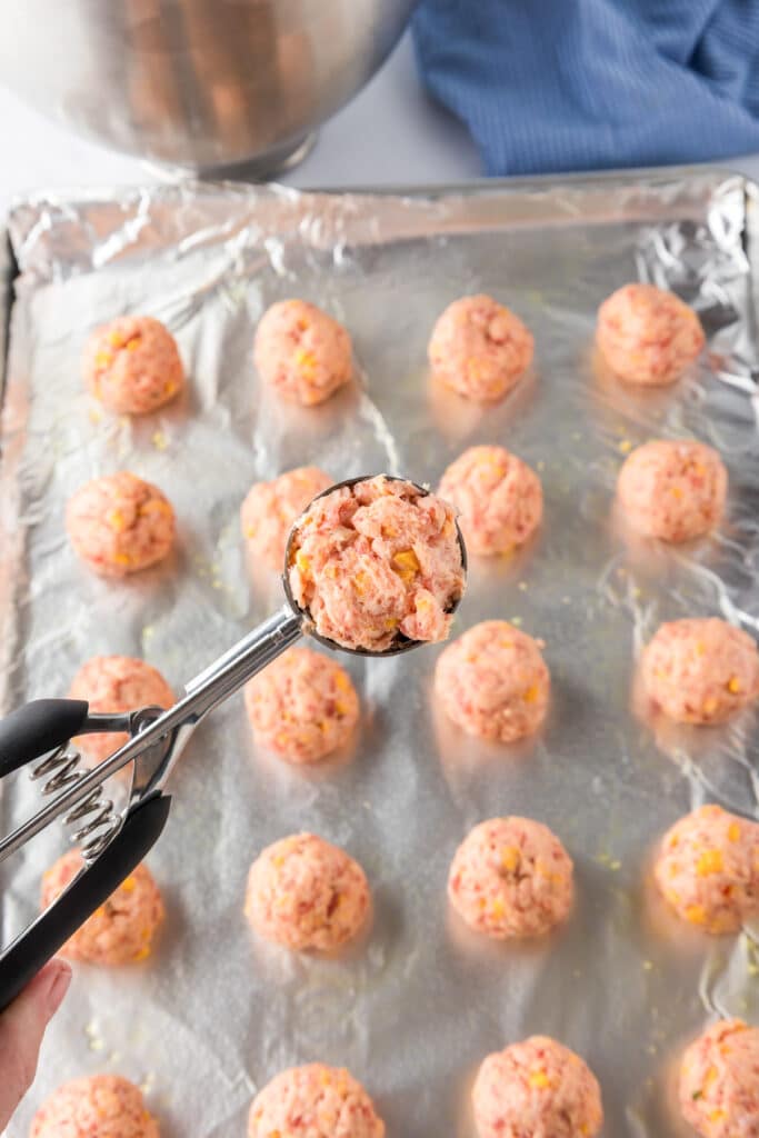 A metal scoop holds a portion of raw cheddar bay sausage balls over a foil lined pan full of more portions.