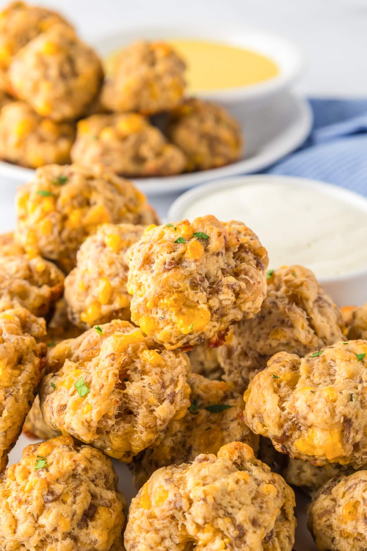 A close-up of cheddar bay sausage balls piled high and close up on a plate with small bowls of mustard and ranch for dipping nearby.