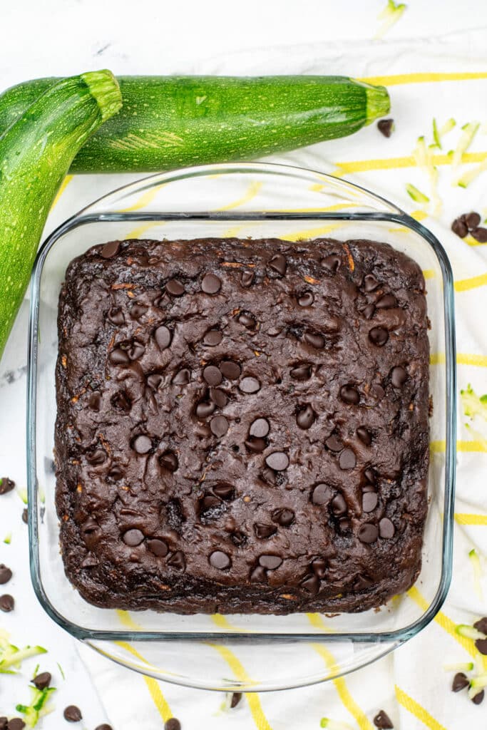 A square glass baking dish with chocolate chip zucchini cake sits on a counter next to two fresh zucchinis.