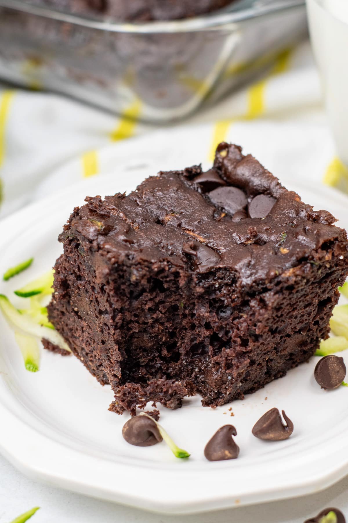 A square slice of chocolate chip zucchini cake on a white plate with extra chips and shredded zucchini.