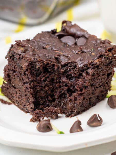 A square piece of chocolate zucchini cake with chocolate chips missing a bite on a plate with chocolate chips on top.