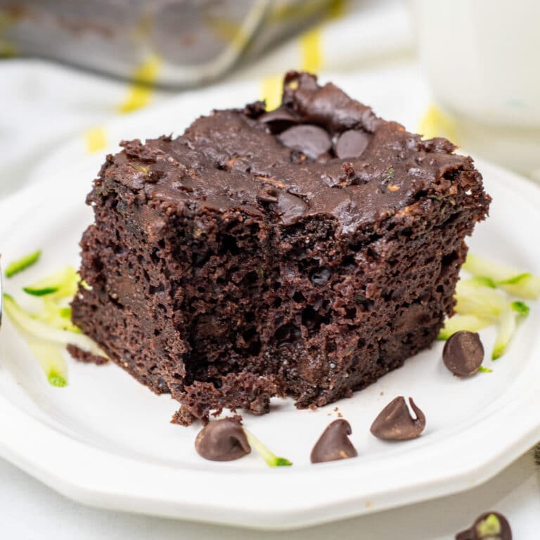 A square piece of chocolate zucchini cake with chocolate chips missing a bite on a plate with chocolate chips on top.
