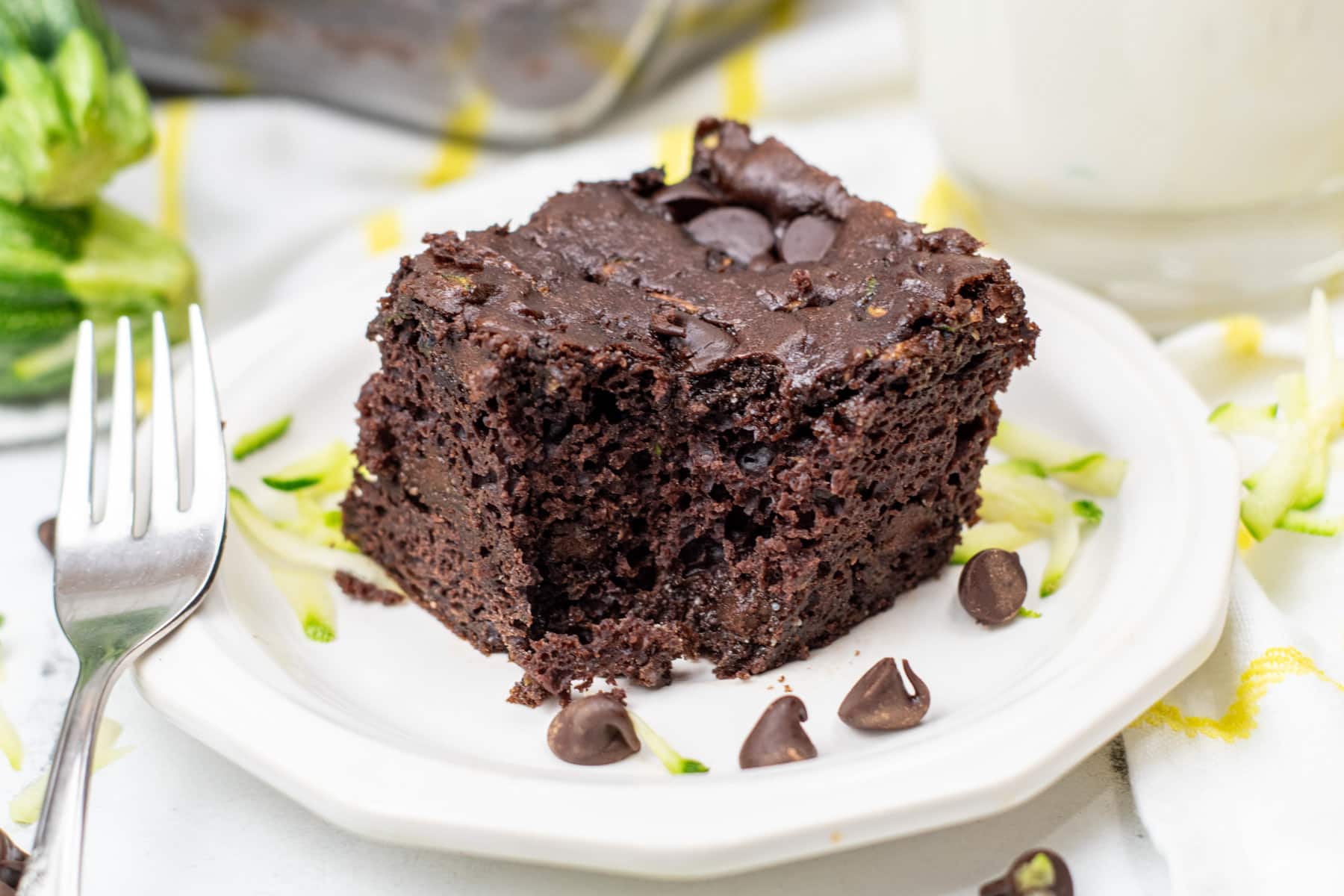 wide view of a piece of chocolate zucchini cake with chocolate chips on a white plate, garnished with grated zucchini, next to a fork.
