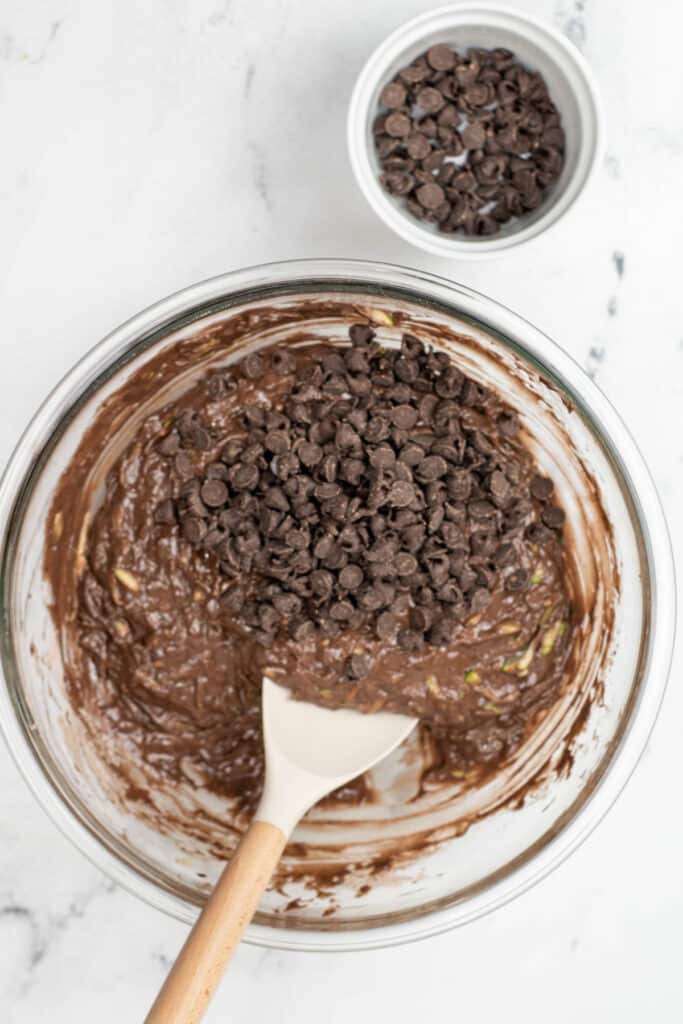 A glass bowl of chocolate batter with chocolate chips and a spatula, next to a small bowl of chocolate chips on a white surface.