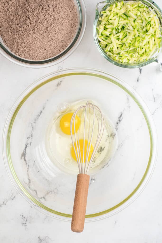A glass bowl with two eggs and a whisk, next to bowls of shredded zucchini and cocoa powder, on a marble countertop.