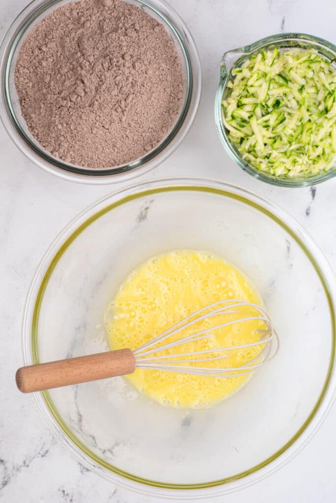 Three bowls on a white surface: one with whisked eggs and a whisk, one with shredded zucchini, and one with dry chocolate cake mix.