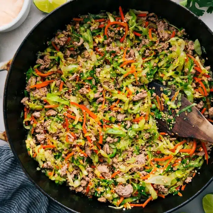 A skillet filled with cooked ground turkey, shredded carrots, cabbage, and green onions, garnished with sesame seeds, next to lime wedges.