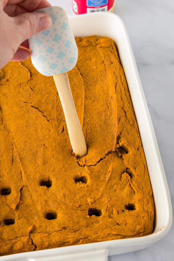 A hand uses a spatula handle to poke holes in a baked pumpkin poke cake in a rectangular baking dish.