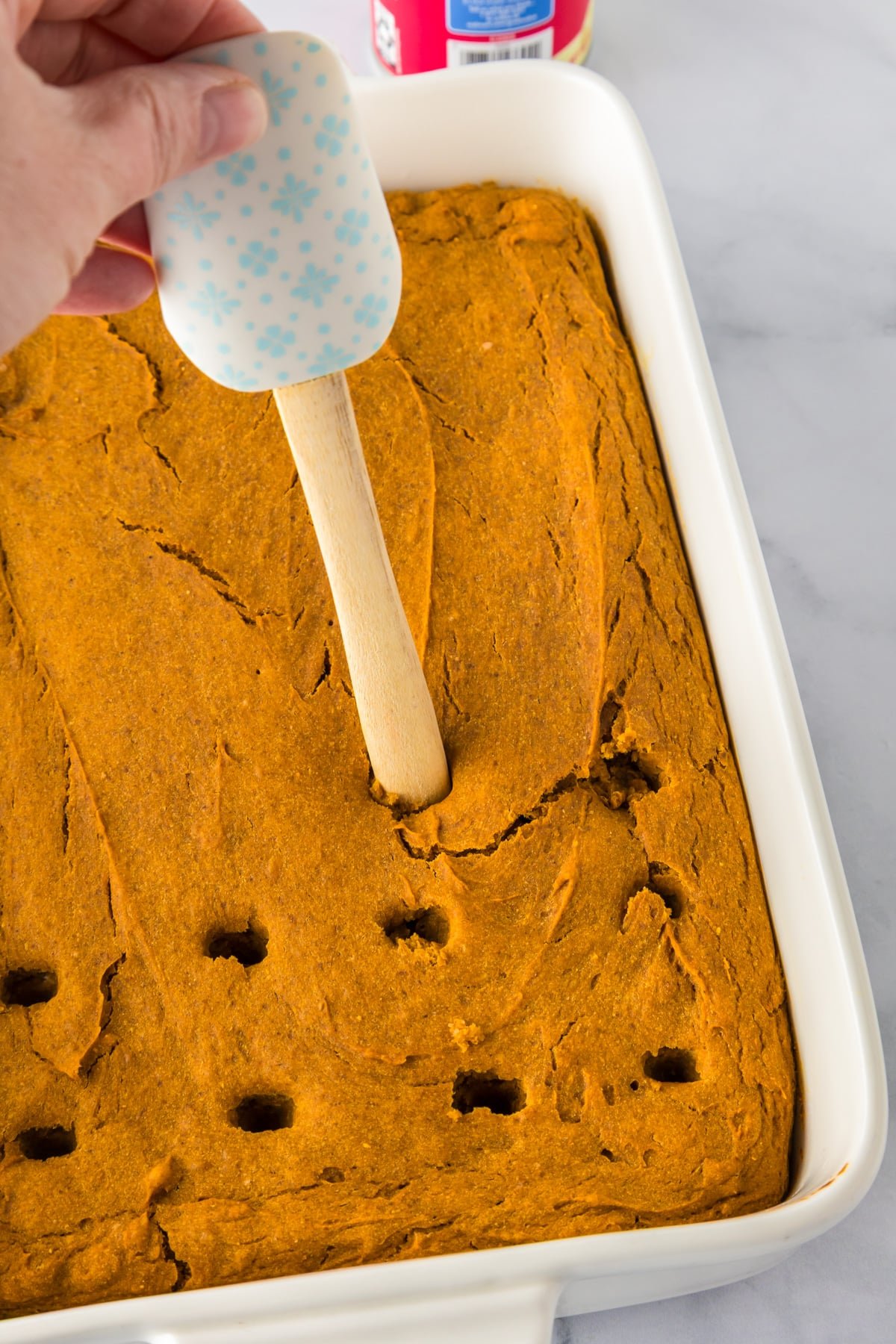 A hand uses a spatula handle to poke holes in a baked pumpkin poke cake in a rectangular baking dish.