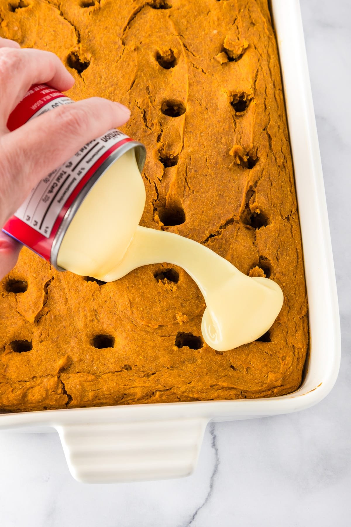 A hand pours sweetened condensed milk from a can onto a baked pumpkin poke cake with holes in a rectangular baking dish.