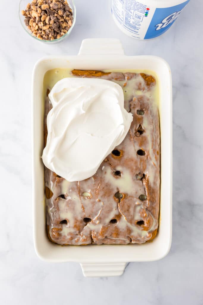 A rectangular baking dish with a pumpkin poke cake soaked in sweetened condensed milk, partially topped with a layer of whipped topping. A bowl of toffee and a whipped topping container are nearby on the counter.