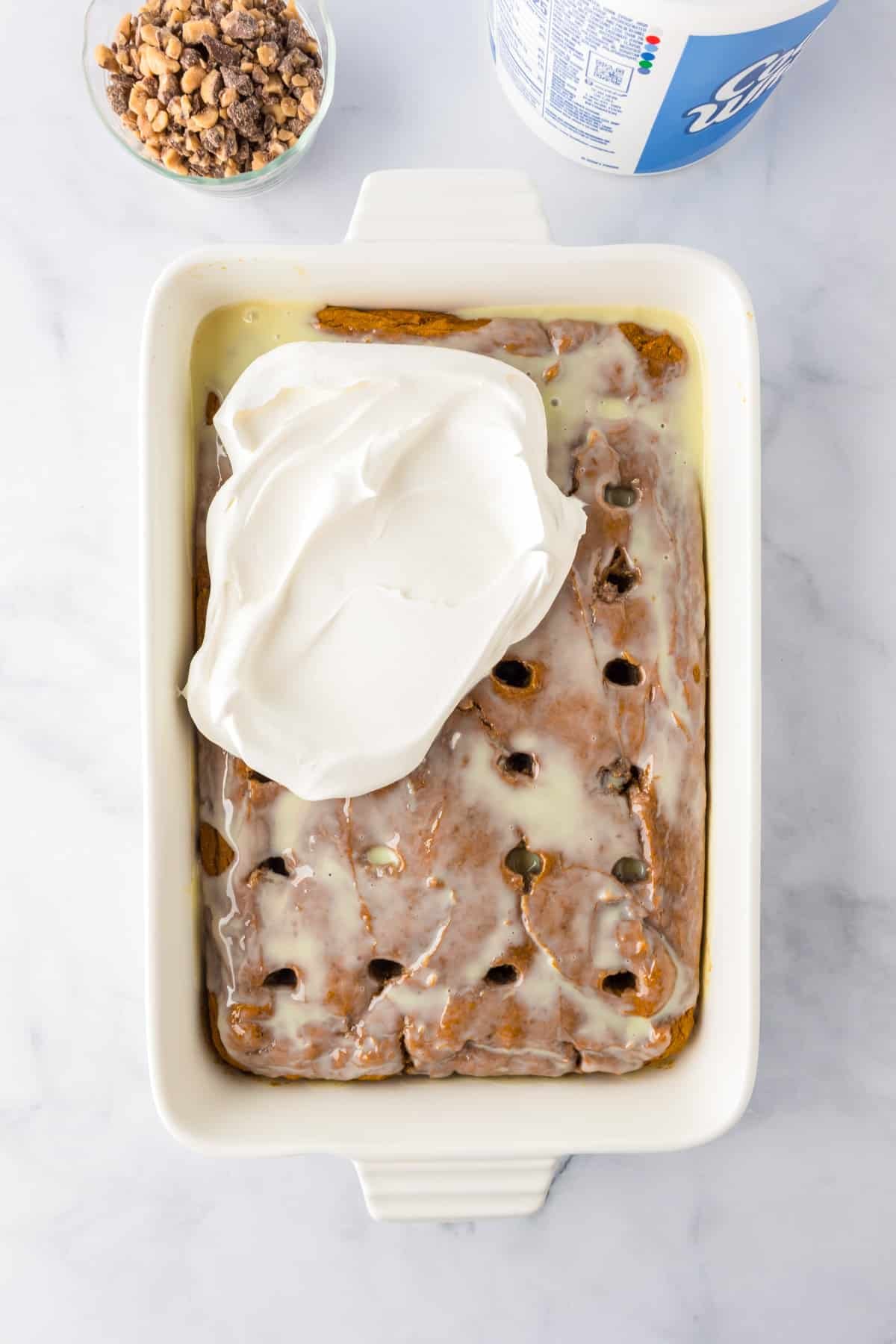 A rectangular baking dish with a pumpkin poke cake soaked in sweetened condensed milk, partially topped with a layer of whipped topping. A bowl of toffee and a whipped topping container are nearby on the counter.