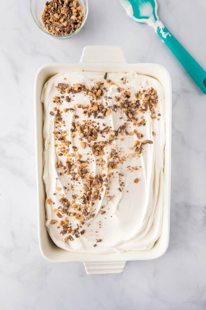 A rectangular dish filled with a pumpkin poke cake being topped with crushed toffee pieces over a whipped topping frosting. A bowl of extra toffee bits and a spatula are nearby on the counter.