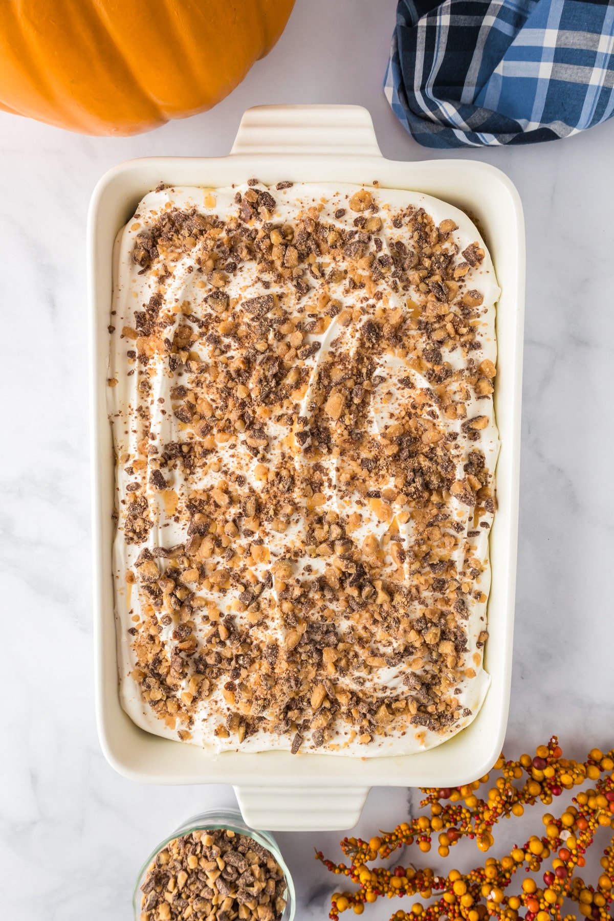 Rectangular dish with a pumpkin poke cake dessert topped with whipped topping and crushed toffee bits on a counter.