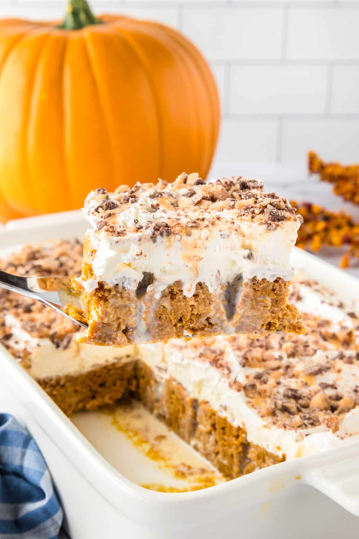 A spatula lifts a slice of pumpkin poke cake dessert topped with whipped topping and crushed toffee chocolate candy from a baking dish, with a pumpkin in the background.