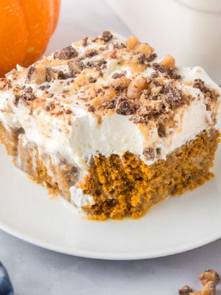 A square slice of pumpkin poke cake with a bite missing, a thick layer of whipped topping and toffee bits are sprinkled on top.