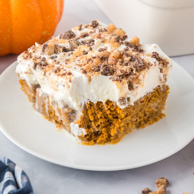 A square slice of pumpkin poke cake with a bite missing, a thick layer of whipped topping and toffee bits are sprinkled on top.