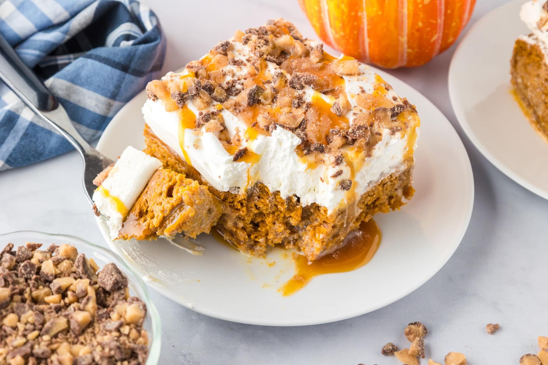 A slice of pumpkin poke cake dessert topped with whipped topping, caramel sauce, and chopped toffee bits sits on a white plate with a forkful taken out.