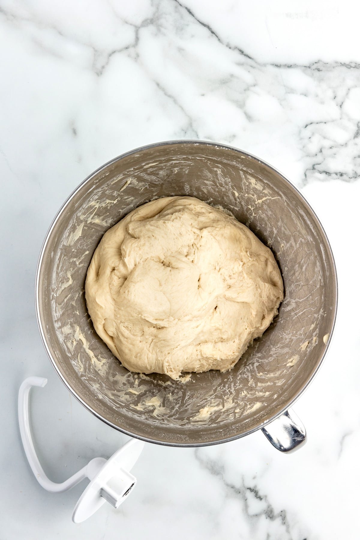 A ball of pizza dough for sheet pan pizza in the base of a stand mixer with a dough hook on the counter nearby.