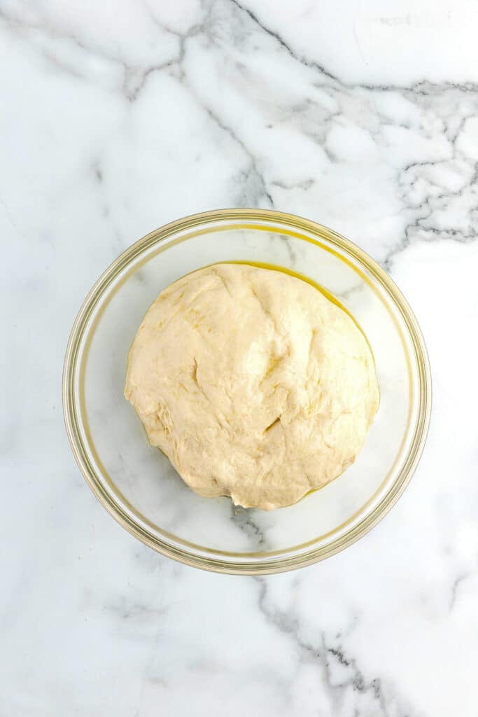 A glass bowl full of pizza dough for sheet pan pizza sits in an oiled bowl.
