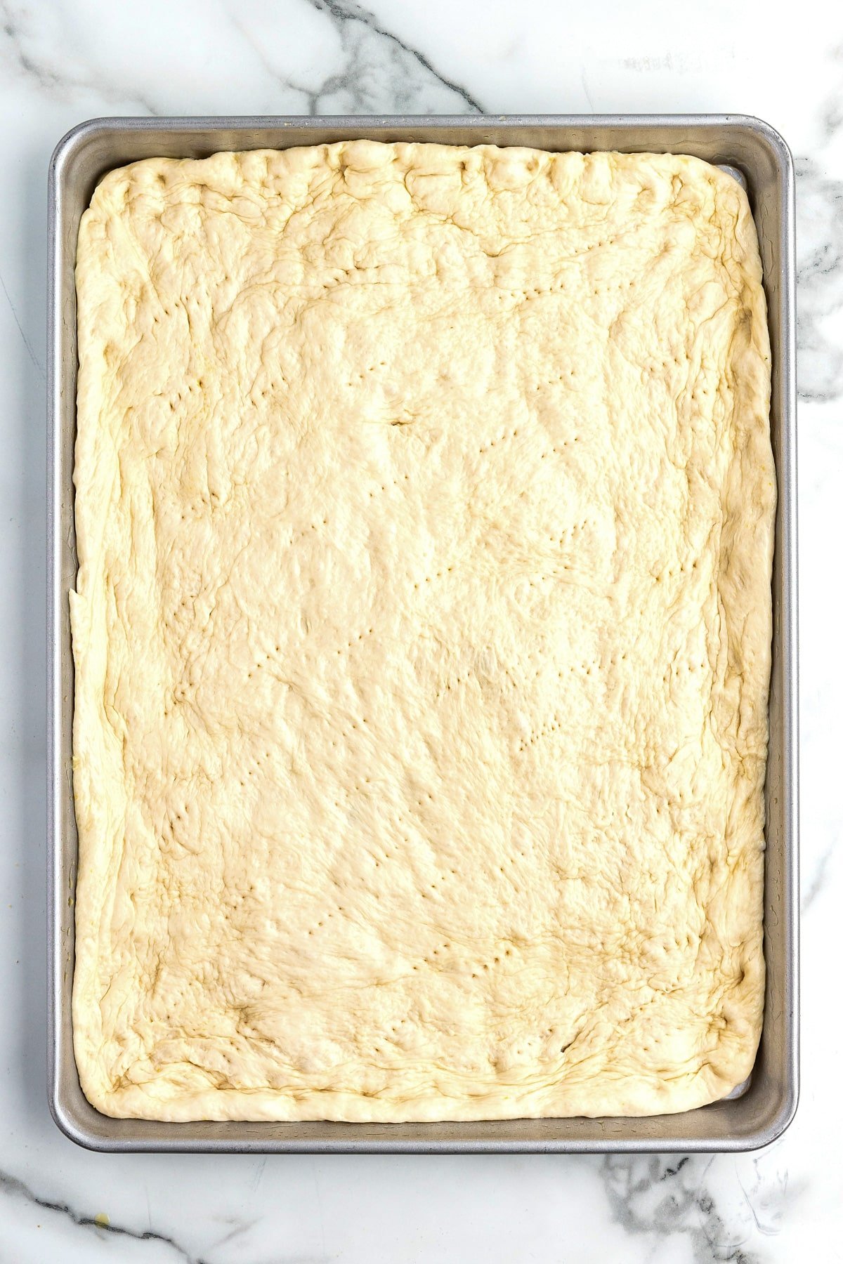 A rectangular sheet pan filled with raw pizza dough for sheetpan pizza evenly spread over the pan with fork pricks all over.