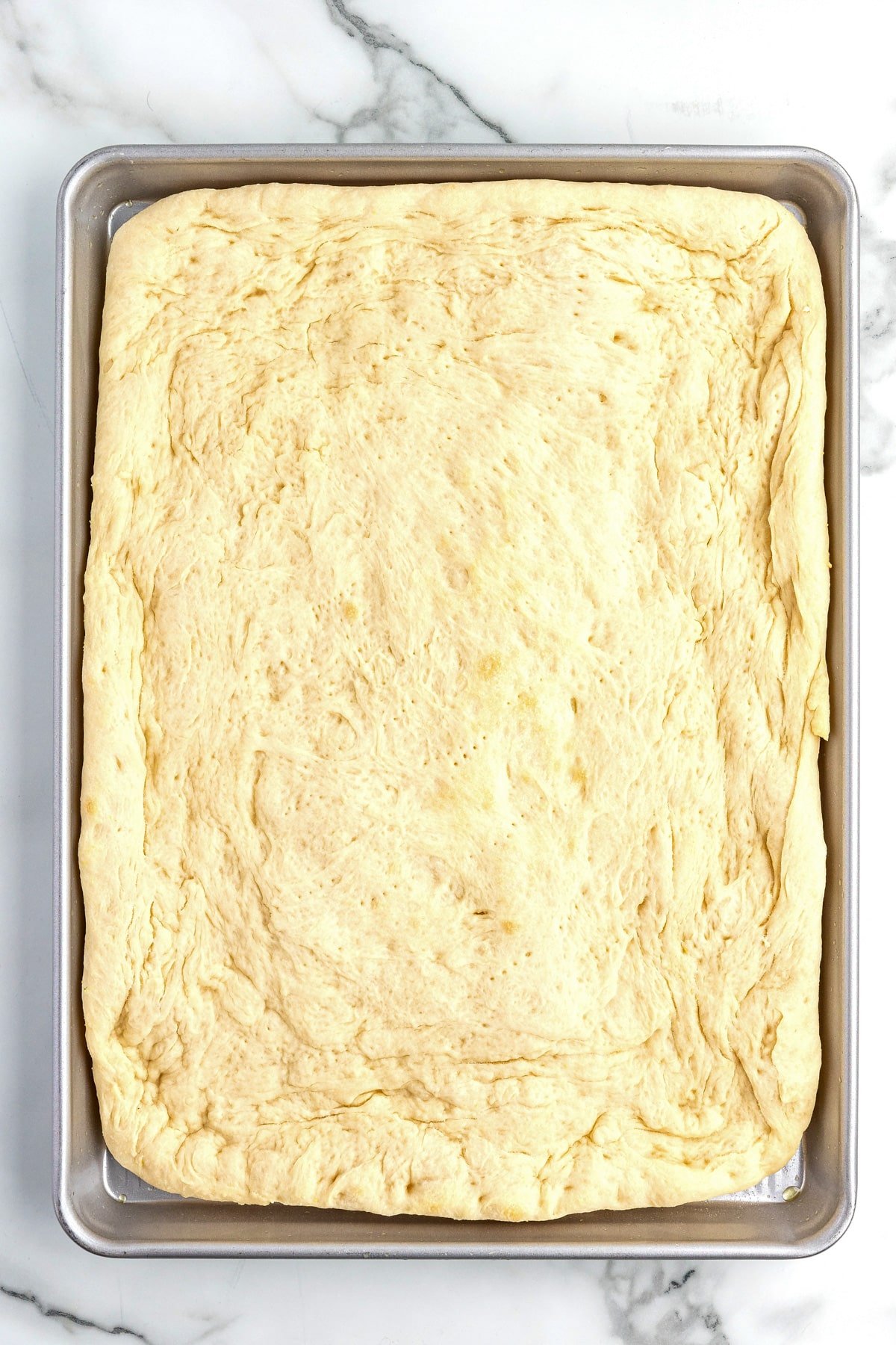 Raw pizza dough spread out on a rectangular metal baking sheet, placed on a white marble surface.