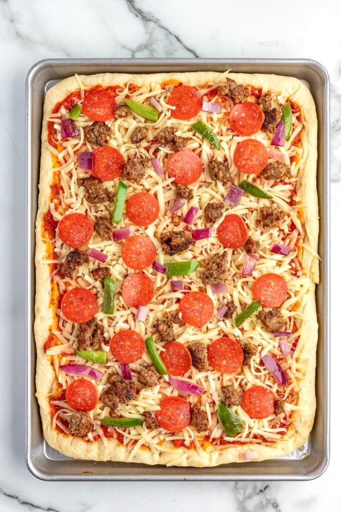Rectangular sheet pan pizza topped with pepperoni, cooked sausage, green bell peppers, red onions, and cheese raw on a counter.