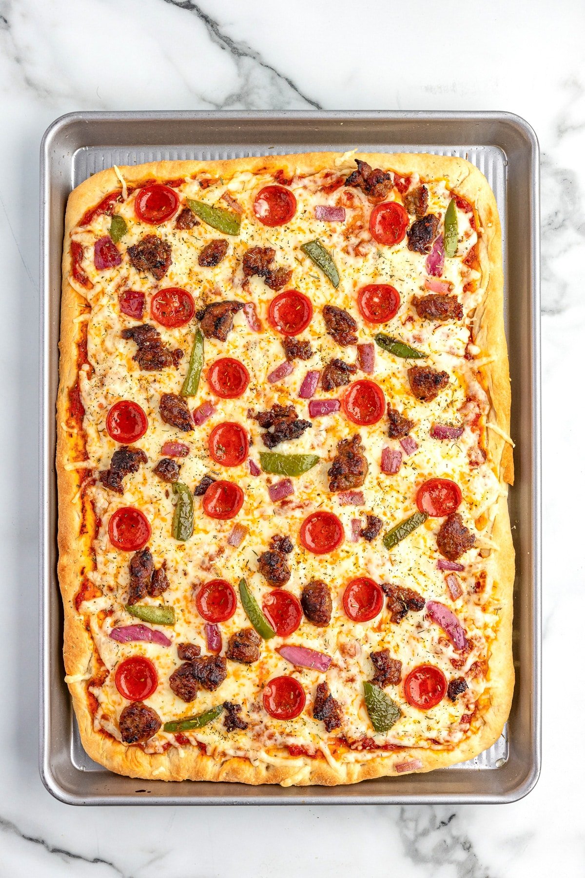 Baked sheet pan pizza on a baking sheet topped with pepperoni, sausage, green bell peppers, and red onions.