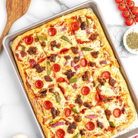Square view of a sheet pan pizza cut into squares topped with cheese, pepperoni, sausage, green peppers, and red onions, placed on a counter with pizza ingredients nearby.