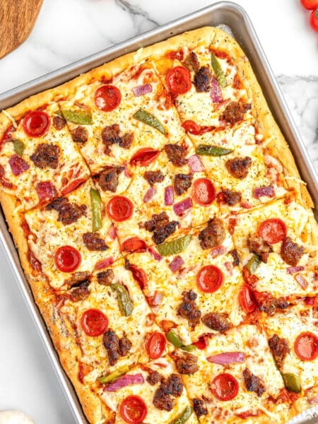 Square view of a sheet pan pizza cut into squares topped with cheese, pepperoni, sausage, green peppers, and red onions, placed on a counter with pizza ingredients nearby.