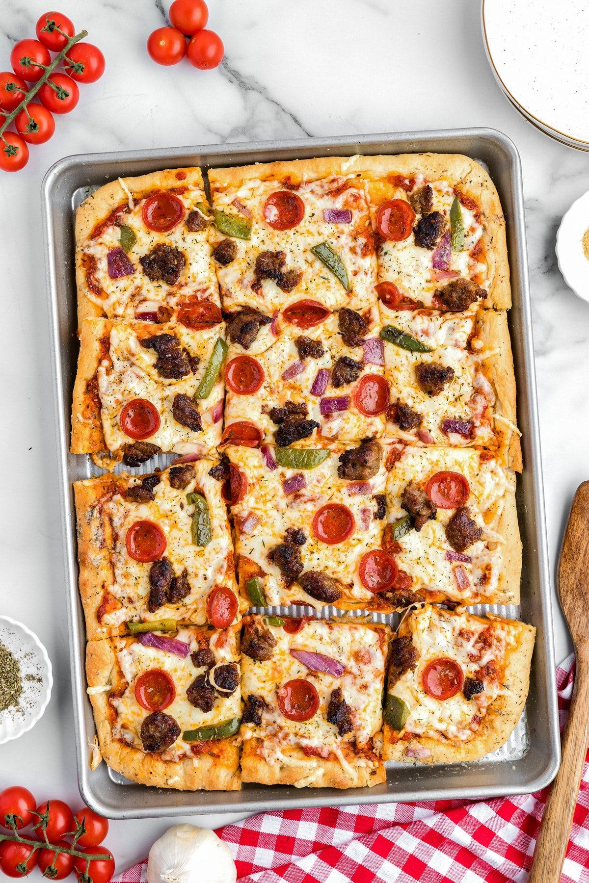 Rectangular sheet pan pizza cut into slices with pepperoni, sausage, bell pepper and onion on a counter.