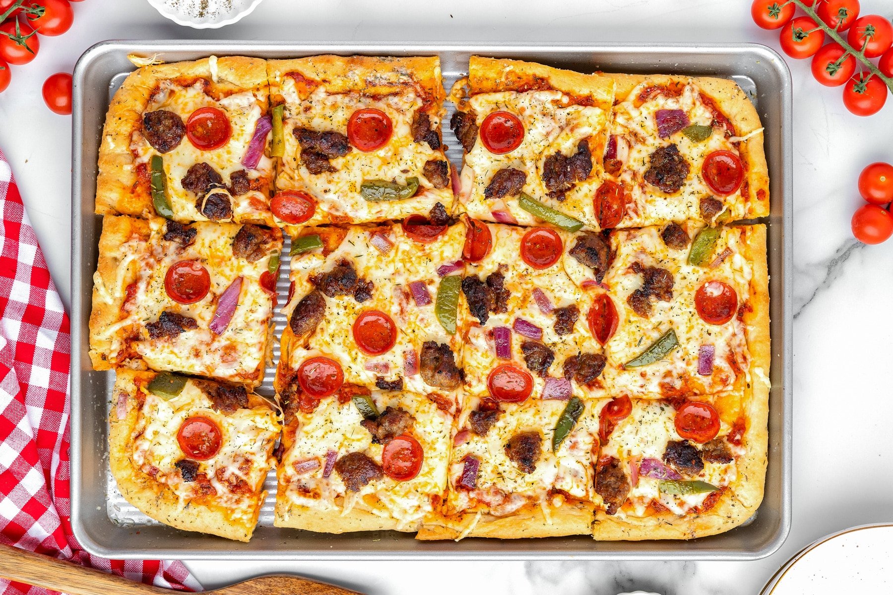 Wide view of rectangular sheet pan pizza cut into 12 square slices, topped with pepperoni, sausage, green peppers, red onions, and cheese.