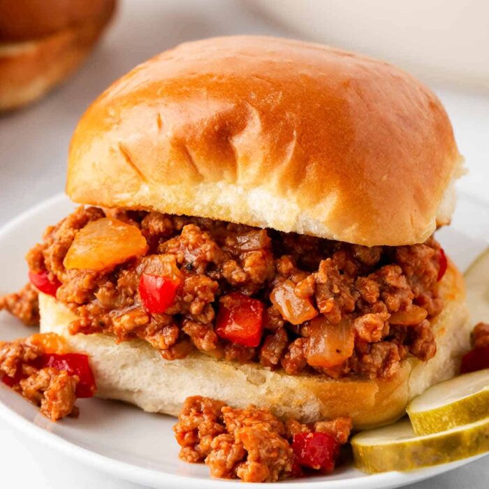A sloppy joe sandwich with ground turkey meat and diced red peppers in sauce, served on a white plate with pickle slices.