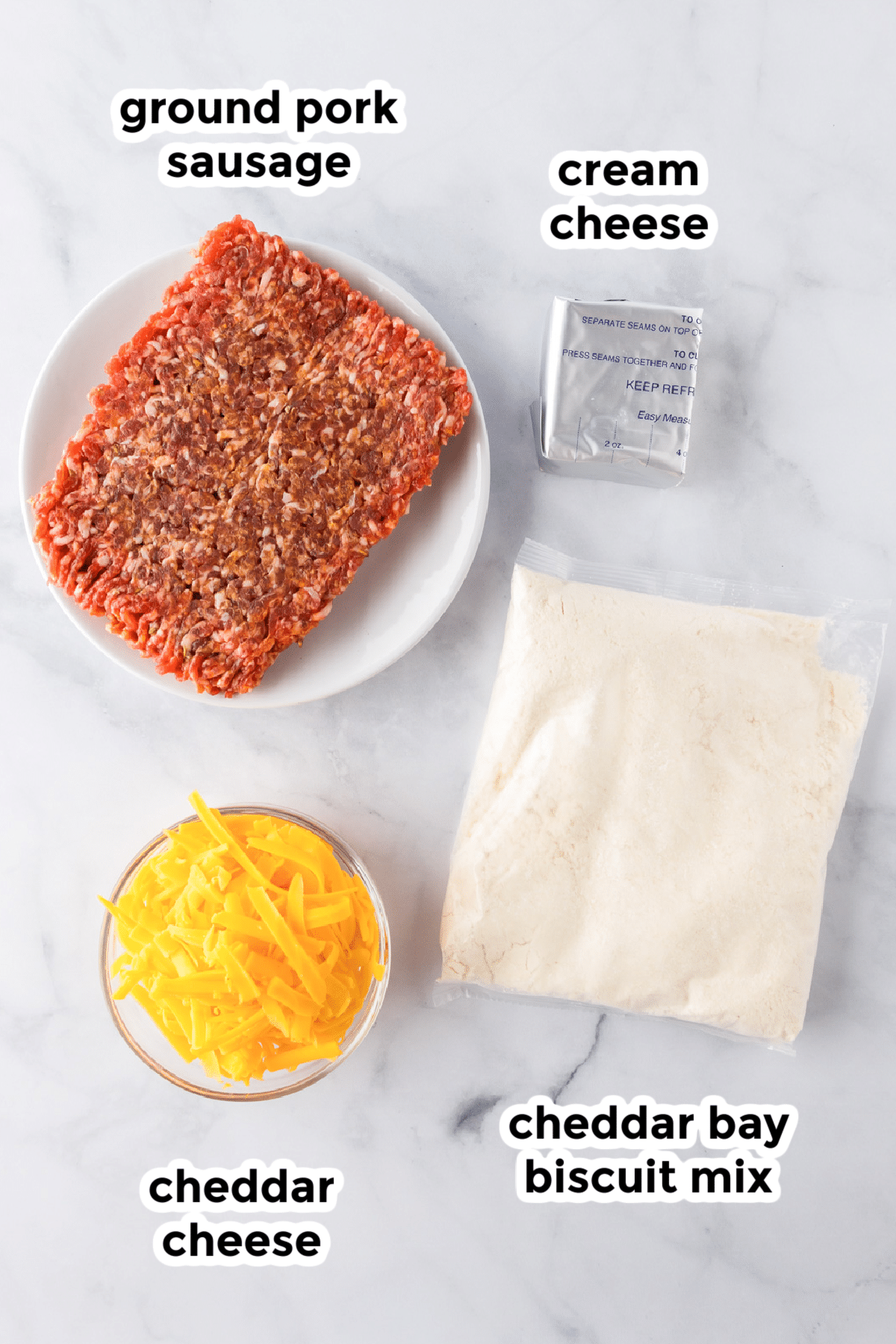 Four labeled ingredients on a countertop: ground pork sausage on a plate, a block of cream cheese, shredded cheddar cheese in a bowl, and a bag of cheddar bay biscuit mix.