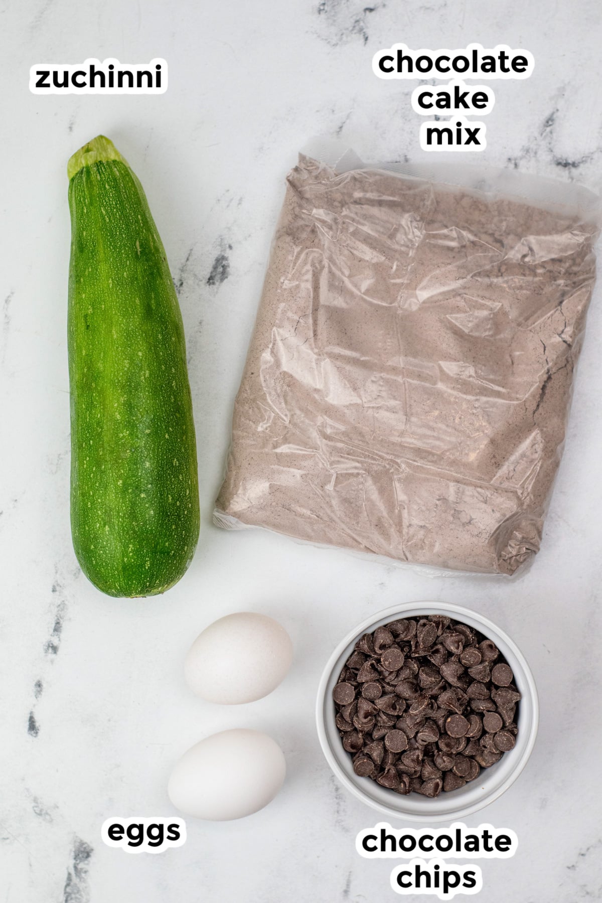 Ingredients for chocolate chip zucchini cake on a counter with text labels.