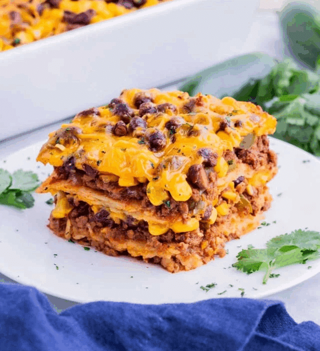 A stack of cheesy Mexican lasagna with black beans, corn, ground meat, and melted cheddar is served on a white plate, garnished with fresh cilantro.