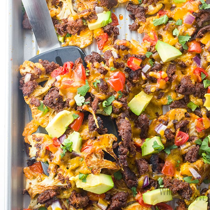 Sheet pan with baked cauliflower nachos with ground turkey topped with melted cheese, avocado, diced tomatoes, red onion, and cilantro; a spatula lifts a portion.