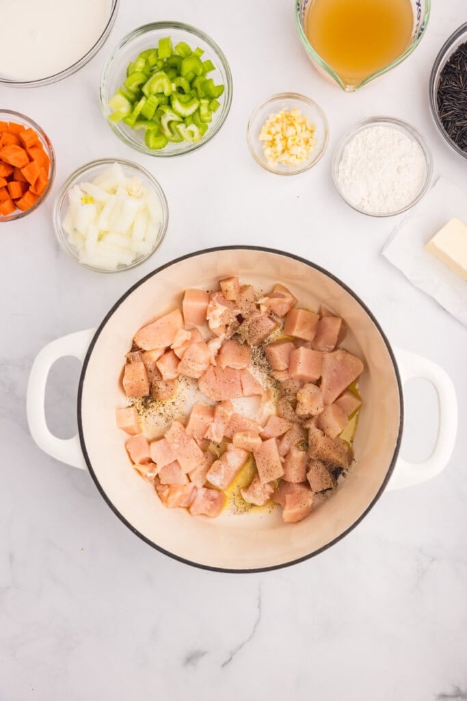 A white pot contains raw, seasoned diced chicken with other ingredients for creamy chicken wild rice soup on a counter.