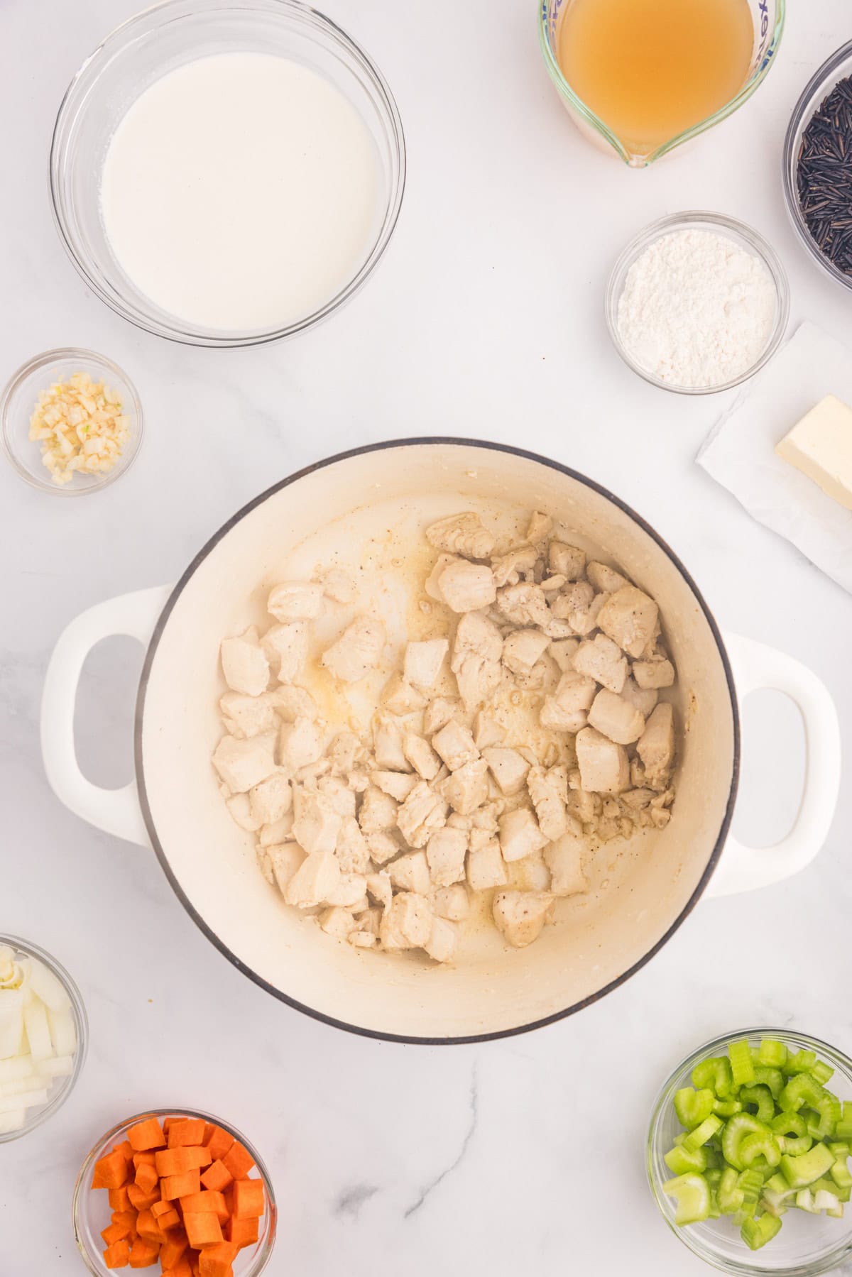 A white pot with cooked chicken pieces for creamy chicken wild rice soup with more ingredients nearby.
