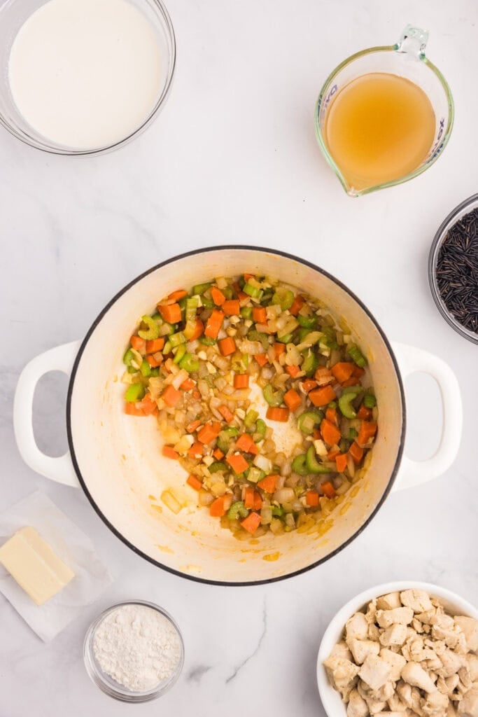 Chopped carrots, celery, and onions are sautéing in a white pot for creamy chicken and wild rice soup surrounded by bowls of flour, butter, broth and cooked chicken.