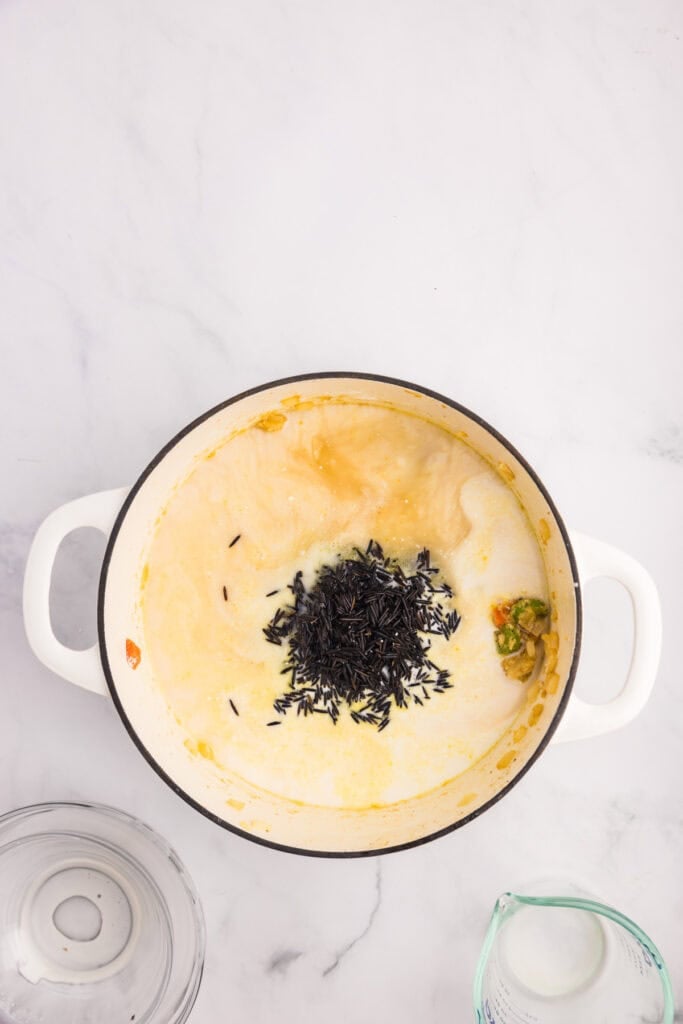 A white pot on a marble surface contains creamy soup with uncooked wild rice and vegetables being cooked to make creamy chicken wild rice soup.