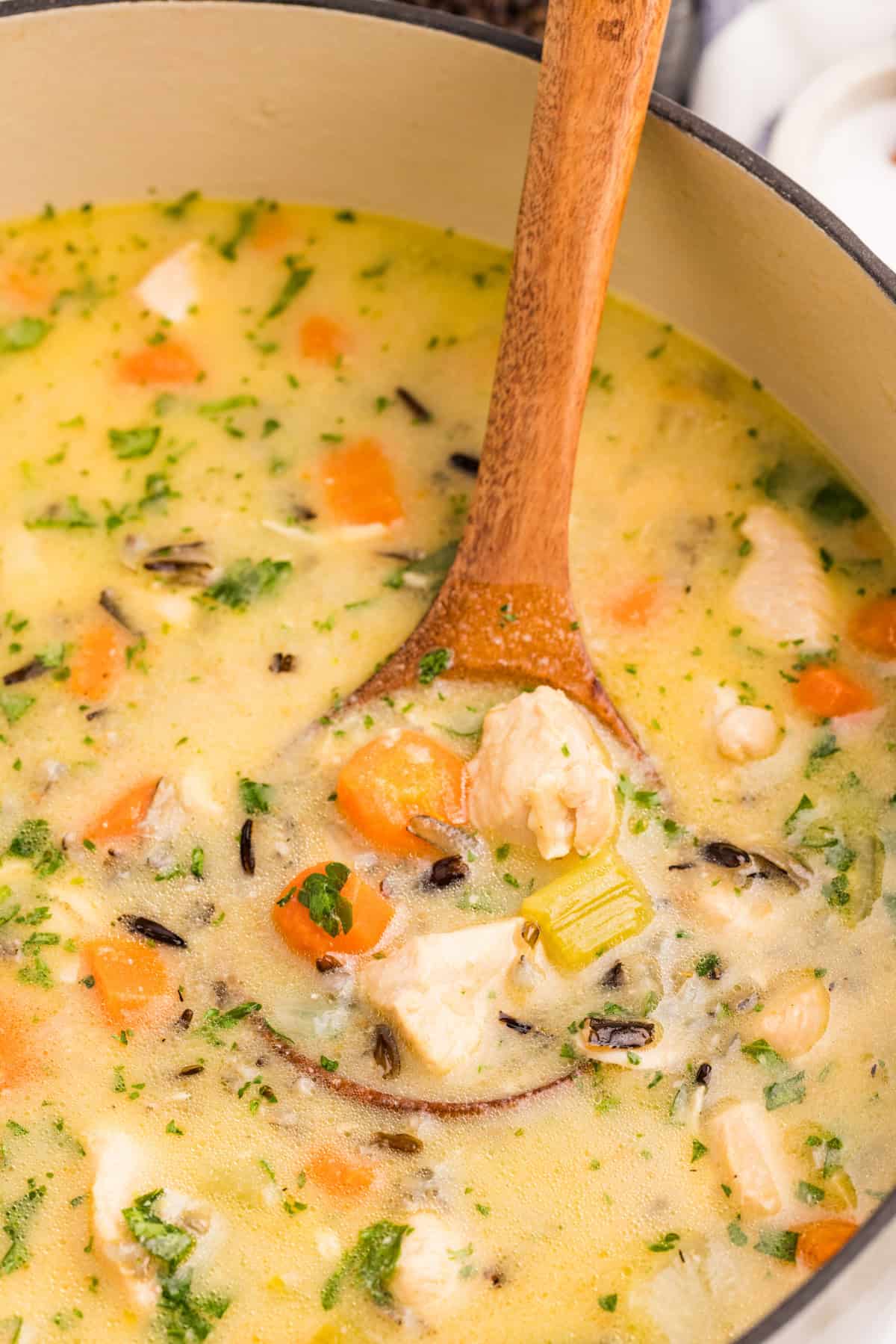 A close-up of a pot of creamy chicken and wild rice soup with carrots, celery, and herbs, being stirred with a wooden spoon.
