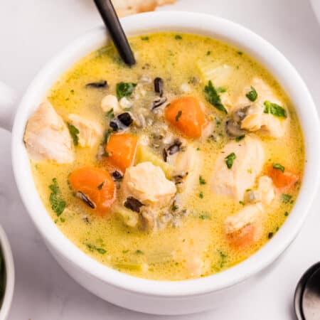 A square view of a bowl of creamy chicken and wild rice soup with chunks of chicken, carrots, and herbs, served with a spoon.