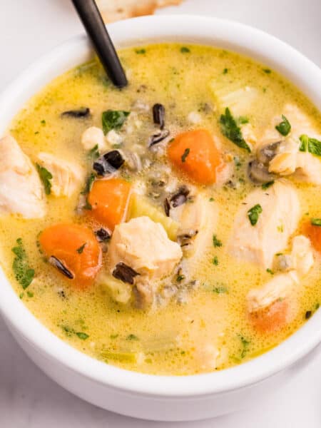 A square view of a bowl of creamy chicken and wild rice soup with chunks of chicken, carrots, and herbs, served with a spoon.