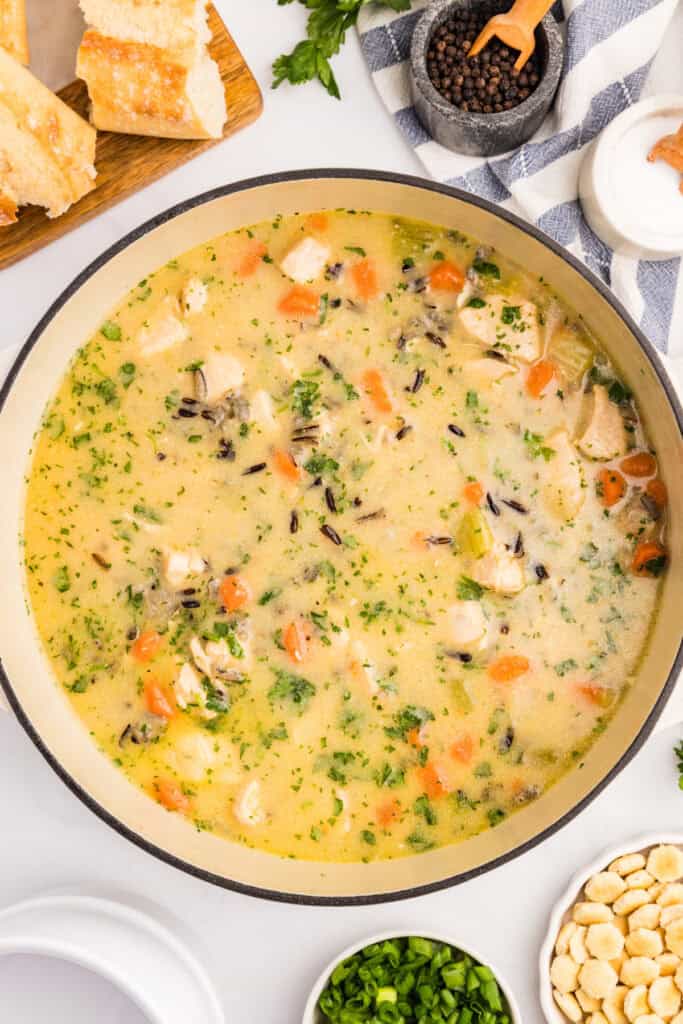A pot of creamy chicken and wild rice soup with chopped vegetables and herbs, surrounded by bread, crackers, and bowls of green onions and seasonings.