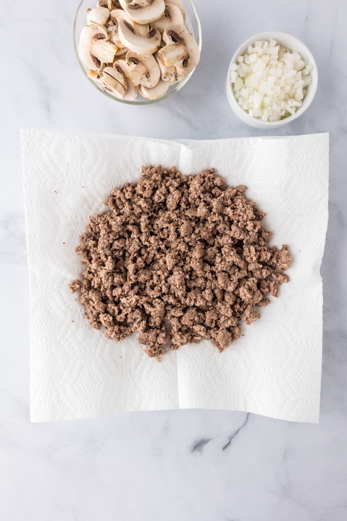 Cooked ground beef draining on paper towels for ground beef stroganoff, with bowls of chopped mushrooms and diced onions on the counter nearby.