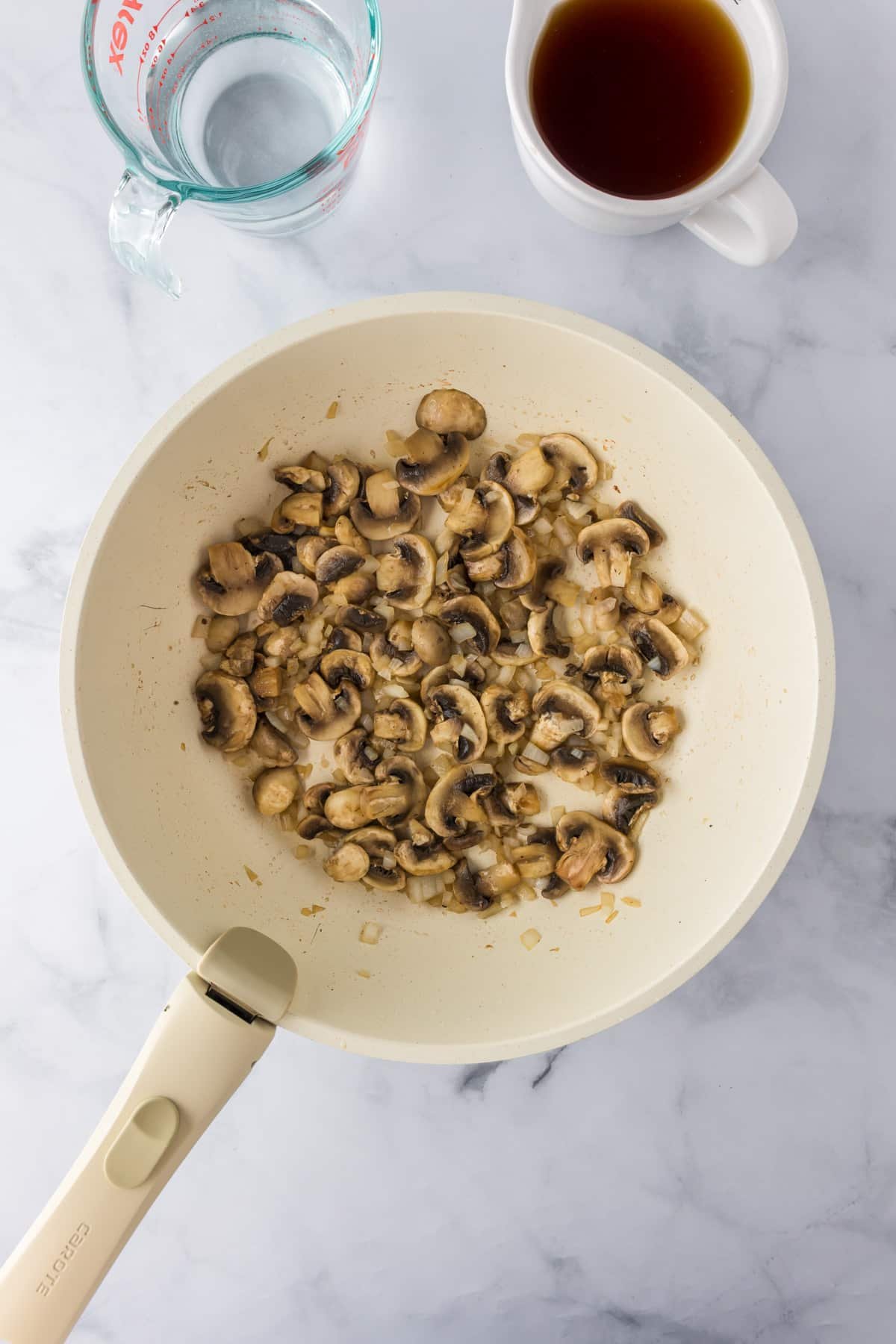 A white skillet with sautéed mushrooms and onions for ground beef stroganoff next to cups of water and beef broth.