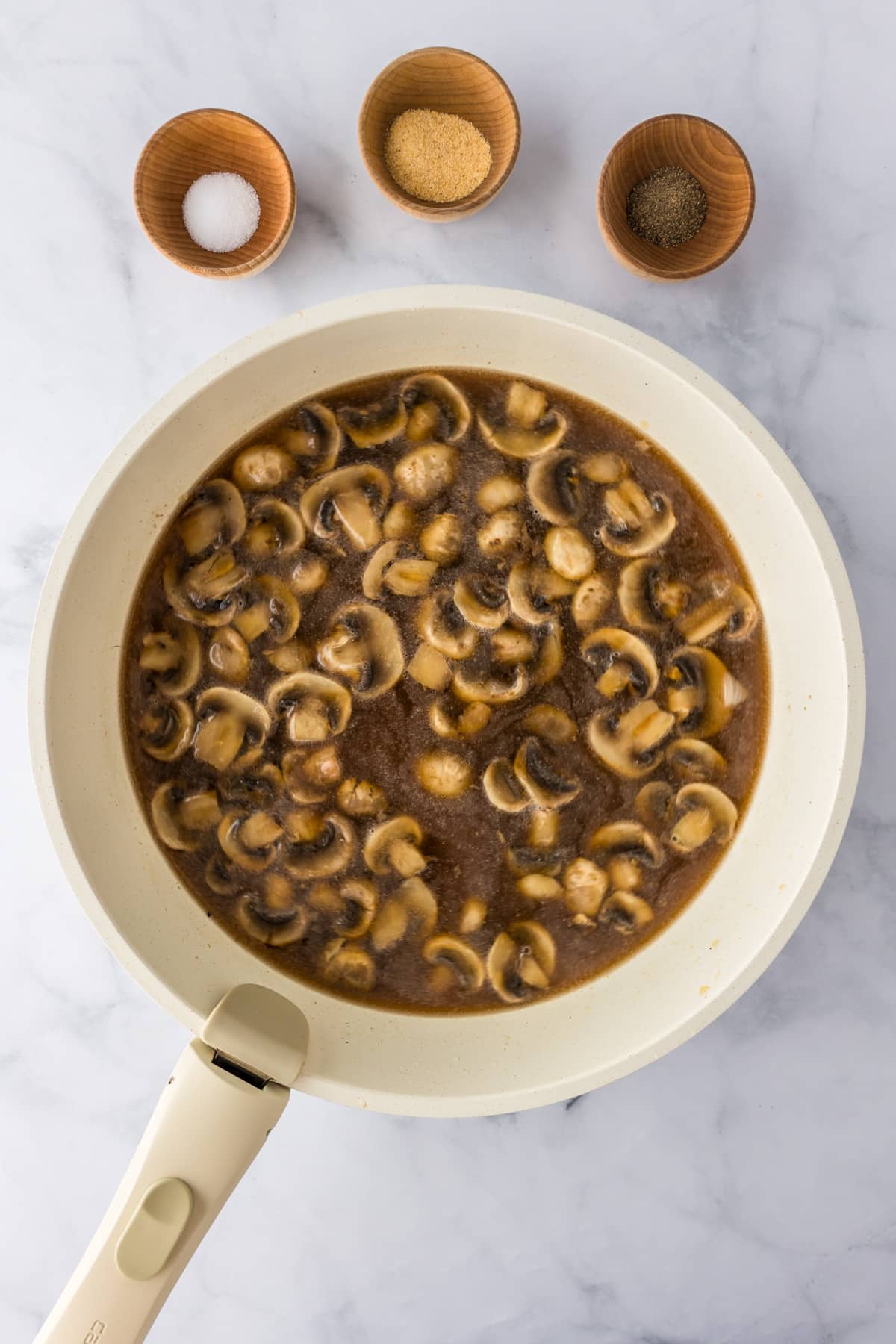 A white skillet with sliced mushrooms cooking in a beef broth gravy for ground beef stroganoff, with salt, pepper and garlic in bowls nearby.