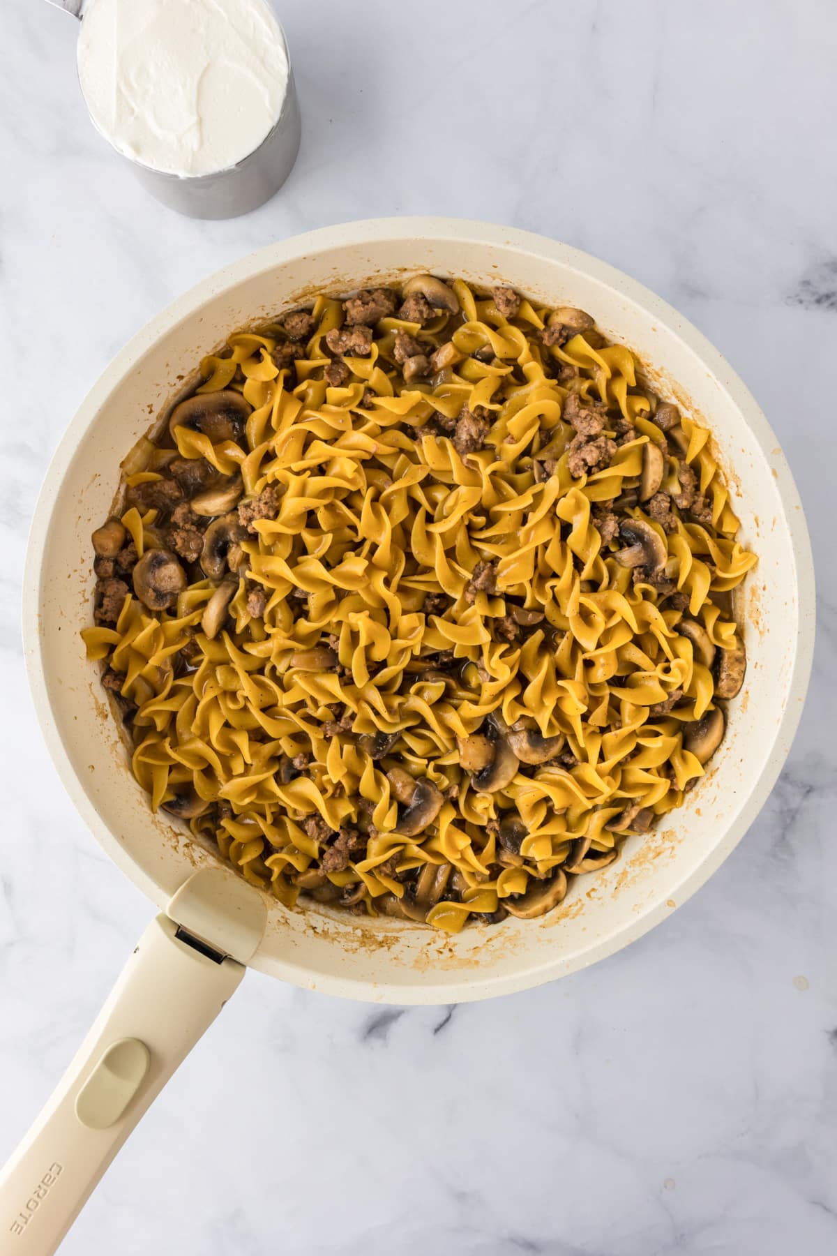 A skillet filled with cooked ground beef stroganoff with sour cream nearby on the counter in a cup.