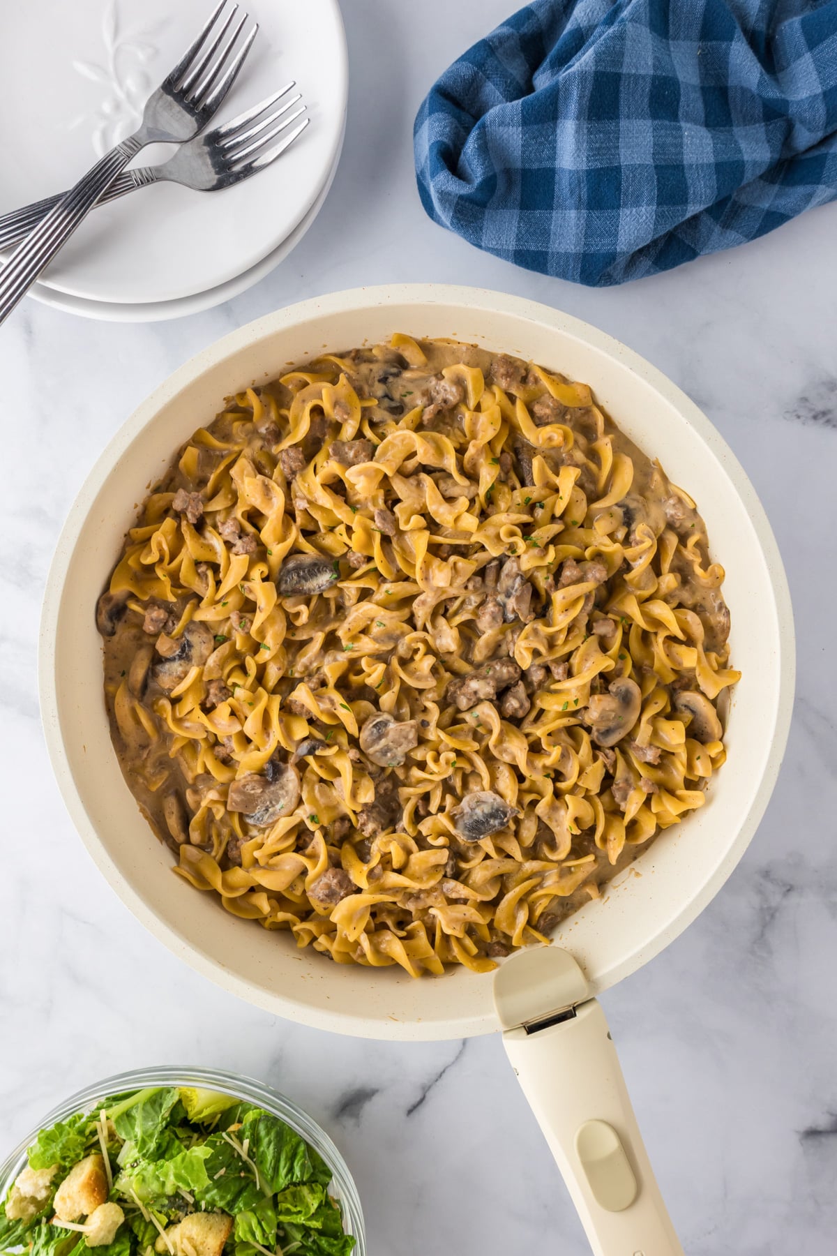 A skillet filled with ground beef stroganoff made with egg noodles and mushrooms on a counter with plates and forks nearby.